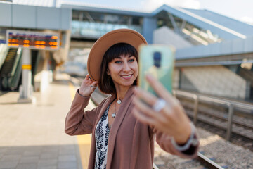 girl waits for a train on the platform and takes a selfie. beautiful tourist woman in a hat takes a selfie at the train station. travel and adventure.
