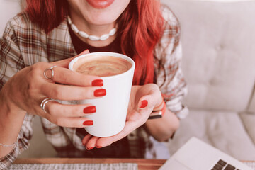 Red haired woman with red manicure holding a warm white coffee mug by her laptop, relaxing in a cozy work from home moment
