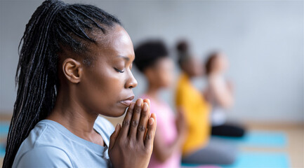 african american woman closing her eyes and holding hands in a prayer pose finding inner peace during a mindful meditation session with other women in a wellness studio