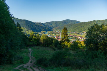 Wide scenic view from a hillside trail overlooking a green valley, distant mountains, and a small town near a winding river. Clear sky and layered forests create a calm travel and adventure mood with 