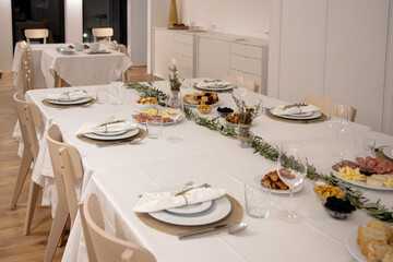 Festive Christmas dinner table in a modern white dining room with long white cloth, plates of pastries, salads, greenery runner, beside lit tree and small tables set for guests during holiday evening.