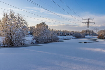 Winter landscape in the Netherlands with a frozen canal, snow-covered trees, and high voltage electricity pylons against a blue sky.
