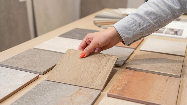 Close-up of a woman's hand with bright orange nails picking a ceramic tile sample from a wooden table. Various stone and clay texture swatches are arranged for an interior decoration project.