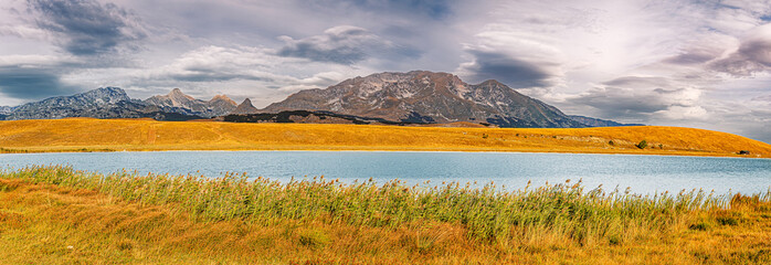Panoramic landscape with a tranquil mountain lake, golden reeds in the foreground, and distant mountains under an overcast sky