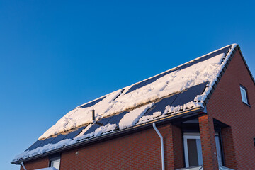 Residential brick house roof with solar panels partially covered in snow against a clear blue sky, reducing energy efficiency.