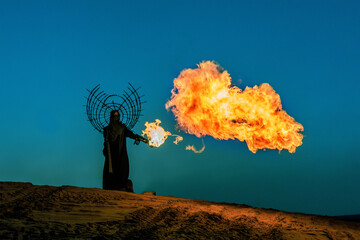 A woman in a cloak and a black crown on her head holds a flamethrower at sunse