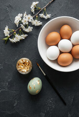 Easter eggs decoration process flat lay. A creative DIY concept with gold leaf, a brush, and cherry blossoms on a dark textured background. Happy Easter!