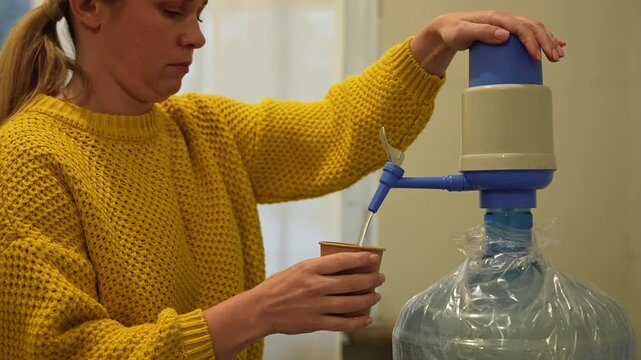 A person in a yellow sweater fills a paper cup with water from a large plastic water dispenser with a manual pump.