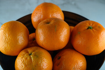 A black bowl holds several fresh oranges arranged in a casual way. The shiny skin of the oranges reflects light in the setting