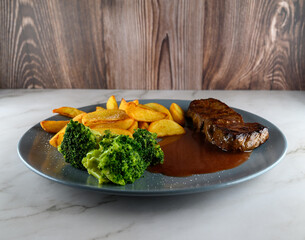 A plate holds slices of steak, golden potato wedges, and bright green broccoli. Brown sauce covers the steak. The setting features a wooden backdrop