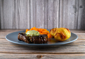Grilled steak sits on a gray plate topped with garlic butter. Sliced carrots and a baked potato accompany the meat on a rustic wooden surface