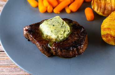 A plate shows a portion of steak topped with a green substance, alongside carrots and an interestingly shaped potato, all set on a wooden table