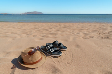 Straw hat, sunglasses, and black sandals on a sandy beach with the seashore in the background