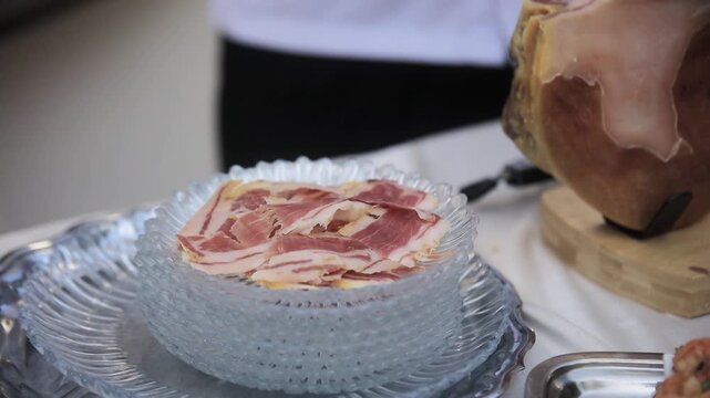 Thin slices of jamon are placed on a glass plate. Thin slices of jamon are placed on a glass plate next to a pork leg. Thin slices of bacon are placed on a glass plate.