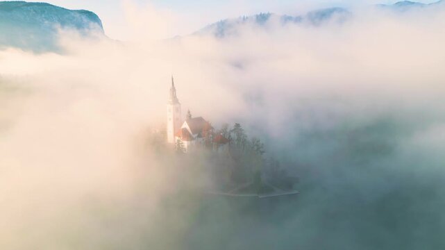 Mari&auml; Himmelfahrt Kirche auf der Insel im See Bled bei Sonnenaufgang, kreisende Kamerabewegung nach oben durch goldenen Nebel, Kirche verschwindet allm&auml;hlich im Dunst im Morgenlicht Slowenien