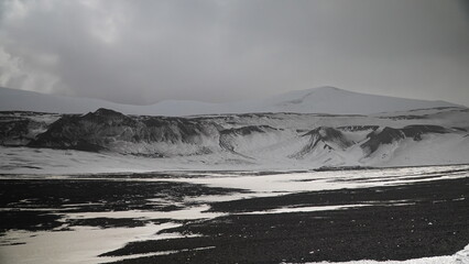 Desolated landscape black volcanic rock mountains lightly covered in snow with a snow mountain backdrop and stormy clouds overhead