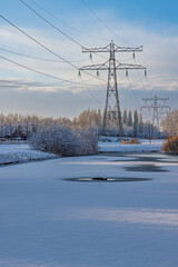 Winter landscape in the Netherlands featuring high voltage power pylons and a frozen canal covered in snow under a cloudy sky.