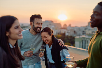 Happy young adults laughing and socializing on a city rooftop during sunset, celebrating with wine glasses