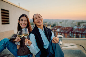 Two diverse women friends toasting with wine glasses, laughing and celebrating on a city rooftop...