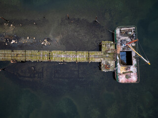 Aerial top-down view of an old pier with a moored barge over dark green water. Industrial marine structure, weathered textures, minimalistic composition, coastal atmosphere