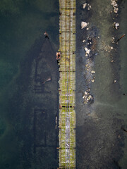 Aerial top-down view of a long wooden pier stretching across shallow coastal water. Weathered structure, rocks beneath the surface, minimalistic geometry, calm marine landscape