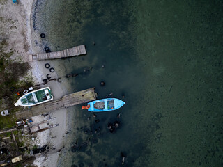 Aerial top-down view of small boats moored at an old wooden pier near a quiet shoreline. Shallow clear water, coastal textures, rustic harbor atmosphere, natural colors and calm seaside mood