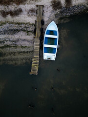 Top-down aerial view of a small white boat moored to a narrow wooden pier near a sandy shoreline. Calm shallow water, minimal composition, rustic coastal atmosphere and quiet seaside mood