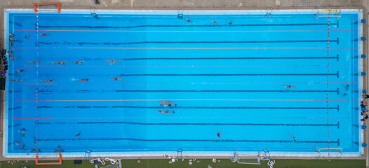 Top-down aerial view of an outdoor swimming pool with multiple swimmers in lanes. Bright blue water, geometric symmetry, sport and recreation concept, summer training atmosphere