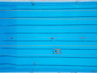 Top-down aerial view of an outdoor swimming pool with multiple swimmers in lanes. Bright blue water, geometric symmetry, sport and recreation concept, summer training atmosphere