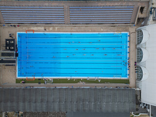 Top-down aerial view of an outdoor swimming pool with multiple swimmers in lanes. Bright blue water, geometric symmetry, sport and recreation concept, summer training atmosphere