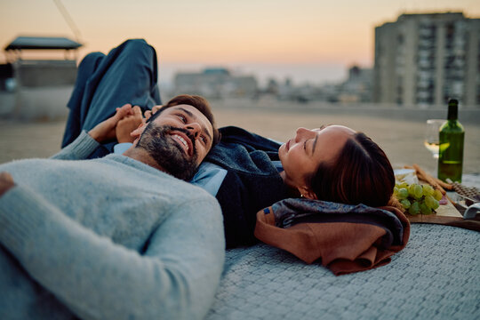 Young couple lying on a blanket, relaxing and enjoying a romantic picnic on a city rooftop at sunset with wine and snacks