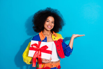 Young fashionable woman with a colorful jacket holds a gift on a blue background smiling and...