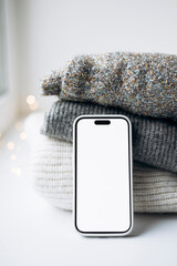 Smartphone with blank screen on cozy knitted sweaters near window.