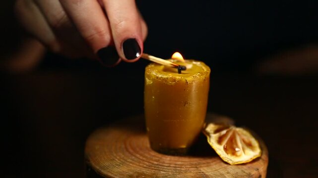 Woman hand lights a match the bees wax candle on wooden candlestick for mood, relaxation, meditation dark black background. Person lighting the decoration handmade single candle flame. Closeup