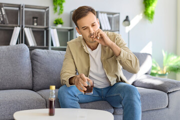 Fototapeta premium Man with alcohol addiction smoking in stress. Sitting on a sofa, he clutches a drink and reflects on problem, signaling poor health, risk, and rising habit. Harmful alcohol habit and risk.