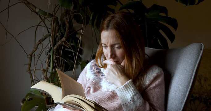 A medium shot of a woman sitting in a grey armchair reading a book titled El infinito en un junco. The setting is domestic with plants.