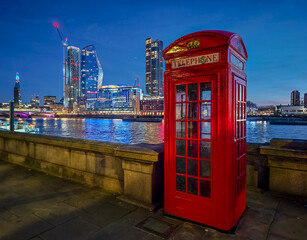Red London telephone booth at night - iconic British landmark glowing in dark urban London street and Thames in illuminated skyscrapers background