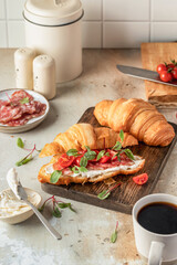 Croissant sandwich with salami, sausage, cherry tomatoes, soft cheese and chard micro greens on wooden board kitchen table background for savory healthy breakfast