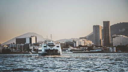 Passenger ferry cruising across rippled bay toward modern city skyline and mountains at sunset, warm golden light, coastal metropolis waterfront, wide sky copy space