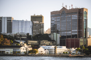 Urban waterfront skyline with modern office towers and glass facades above low historic buildings, calm bay water foreground, warm morning light, business district cityscape