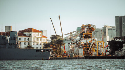 Industrial harbor waterfront with docked ship and huge offshore platform equipment, cranes and shipyard structures beside historic building, modern city skyline in warm light