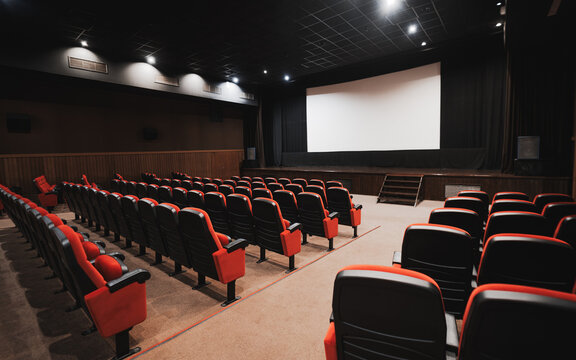 Empty modern movie theater auditorium with rows of red and black seats facing large blank projection screen, stage and steps, dim lighting, entertainment venue interior; low-key wide-angle shot