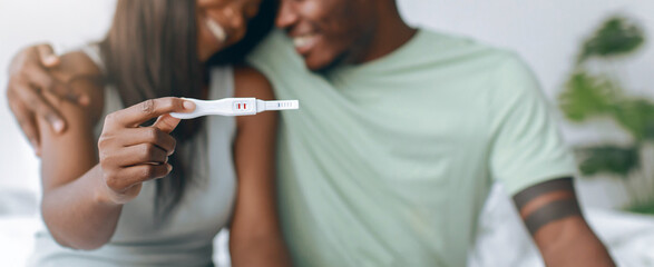 A couple sits together on a bed, smiling as they look at a pregnancy test in the woman's hand. They seem happy and excited about the news.