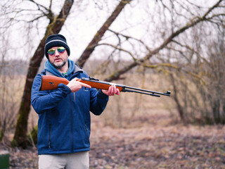 A man in a green jacket and hat is holding a rifle. He is smiling and looks confident handling the weapon. Hunter holding light small caliber gun. Grey winter snowless background.
