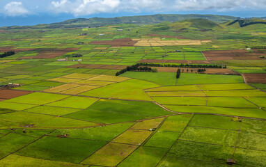 Panoramic view of the colorful agricultural fields from Serra do Cume lookout on Terceira Island, Azores, Portugal. Lush green, yellow, and brown farmlands form a scenic patchwork landscape 