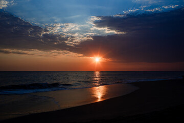 Seascape during sunset featuring dark clouds and golden sunlight reflecting on the wet sand of the shore.