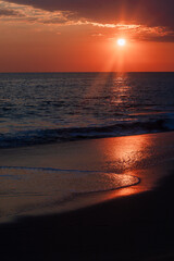 Seascape during sunset featuring dark clouds and golden sunlight reflecting on the wet sand of the shore.