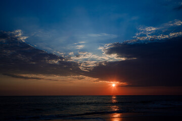 Seascape during sunset featuring dark clouds and golden sunlight reflecting on the wet sand of the shore.