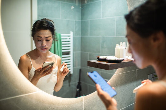 Young adult woman focused on phone while applying face mask in bathroom