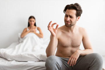 Impotence. Unhappy Man Holding Potency Pill Before Sex While Girlfriend Waiting In Bed Indoors....
